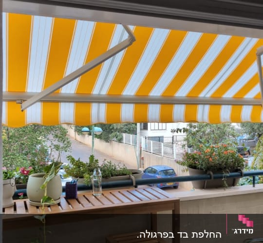 סוכך צהוב לבן מעל מרפסת עם צמחים ורהיטים